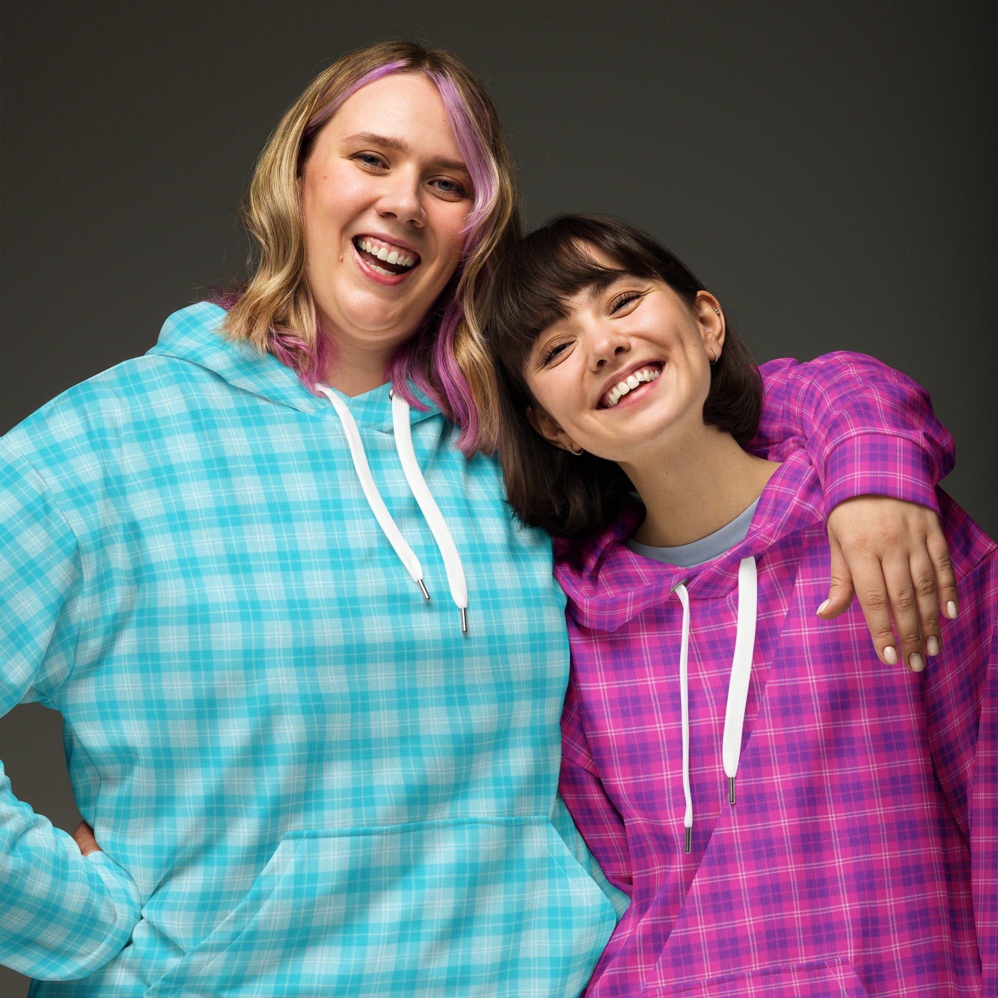 Two individuals wearing vibrant, colorful plaid hoodies made from eco-friendly fabrics. The left hoodie features an electric blue plaid pattern, while the right showcases bold fuchsia and pink plaid.